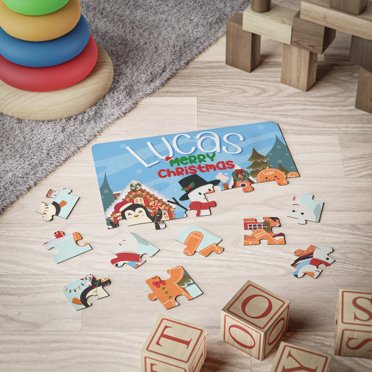 Personalized 30-piece Christmas jigsaw puzzle for kids shown on the floor with scattered pieces, featuring a festive winter village with a snowman, penguin, and gingerbread character, with a customizable child’s name displayed across the top of the puzzle.