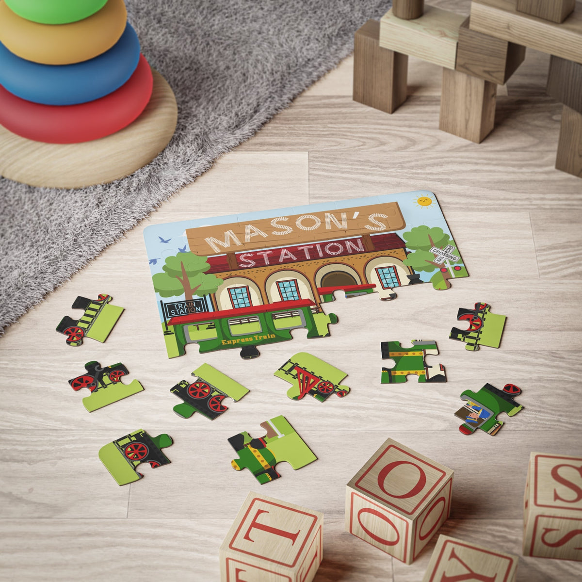 Personalized train station jigsaw puzzle for kids shown in a playroom setting, with jigsaw pieces scattered and a customizable name printed across the top of the puzzle.