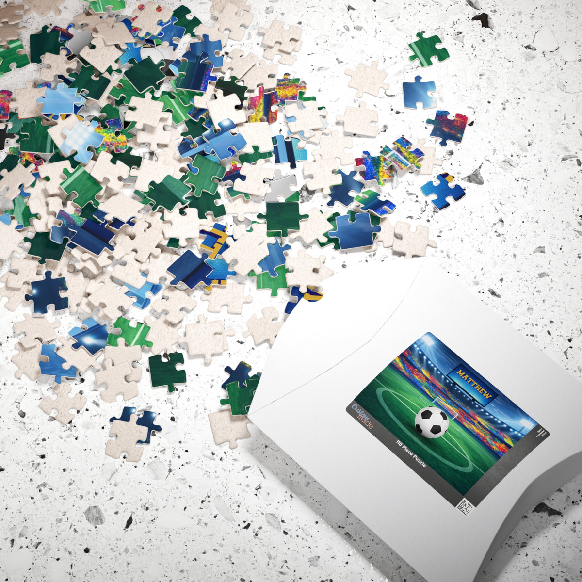 Personalized soccer stadium jigsaw puzzle shown with loose jigsaw pieces spread out next to the puzzle box. Stadium scene includes a customizable name on the jumbo screen and a customizable soccer ball. 110-piece puzzle.