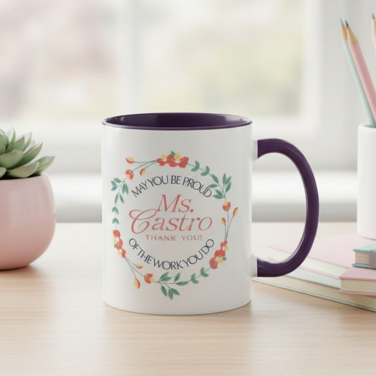 Personalized ceramic accent mug with colored handle and interior displayed on a desk, featuring a floral wreath design with a customizable name printed in the center on the front of the mug and the quote “May You Be Proud of the Work You Do,” styled with notebooks, pencils, and a small plant in the background.