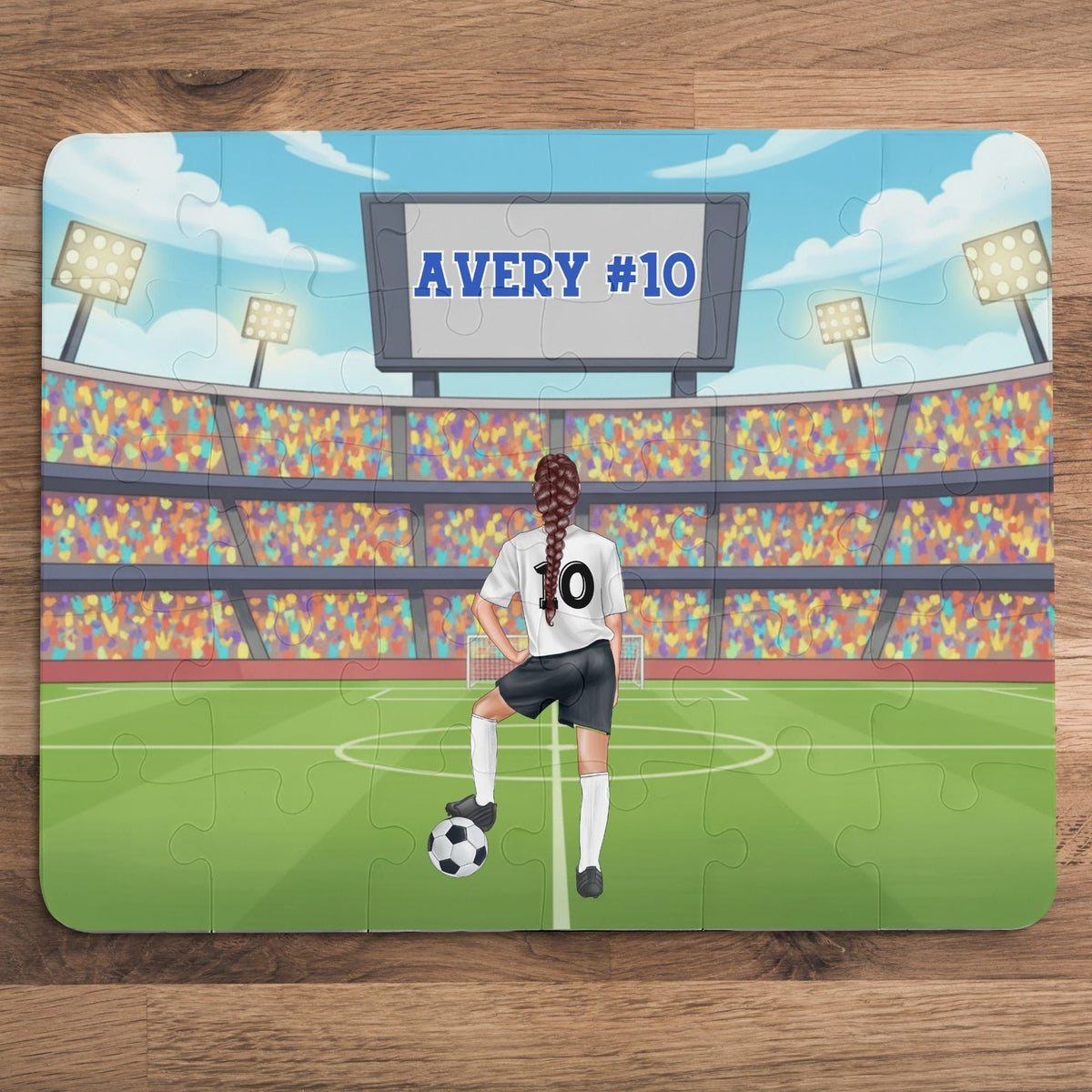 Personalized girls soccer puzzle showing a young player standing on a soccer field inside a large stadium, resting one foot on a soccer ball. The back of the jersey displays a customizable name and number, and a stadium scoreboard above also shows the personalized name and number.