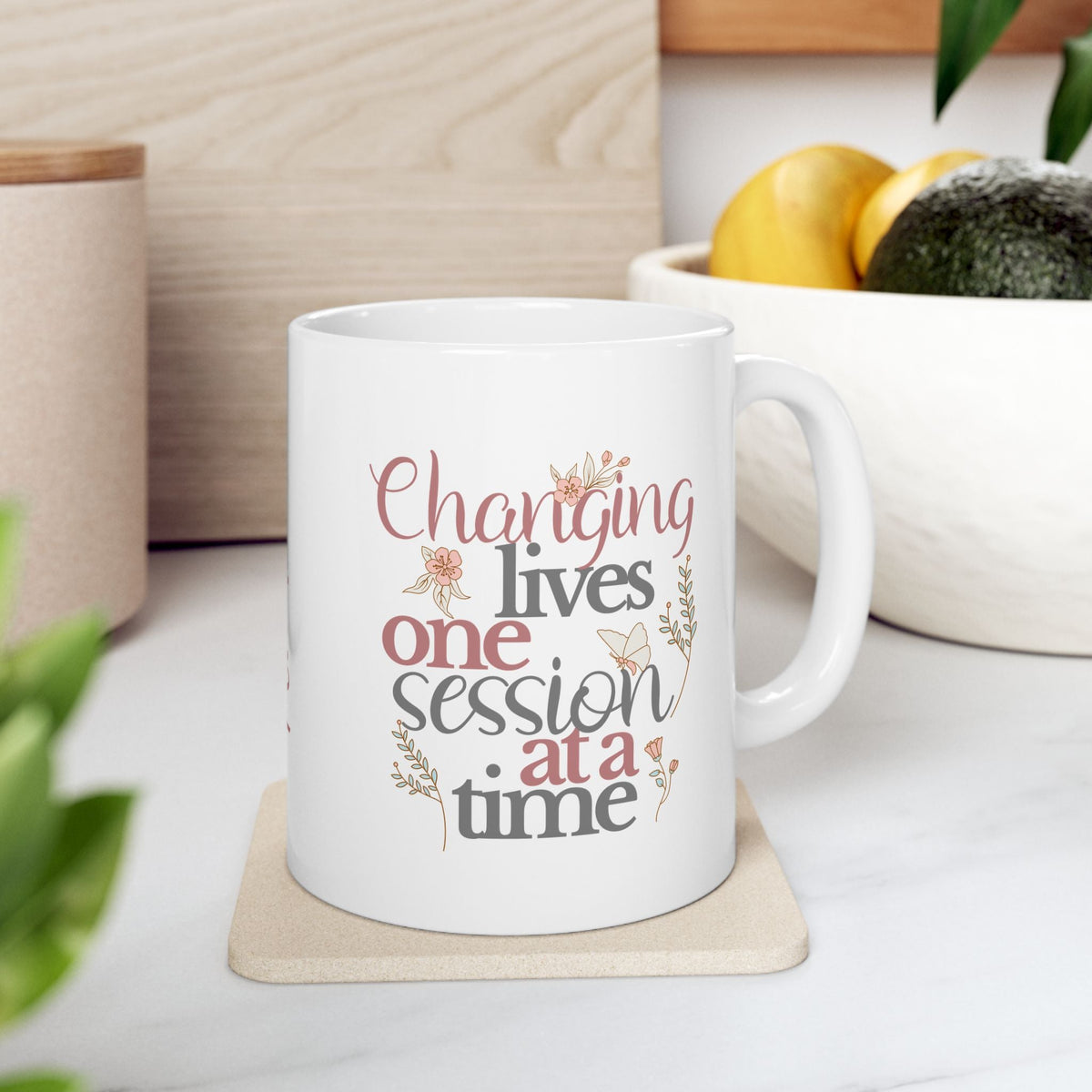 White ceramic mug featuring the phrase “Changing Lives One Session at a Time” in soft floral typography, with a customizable name option printed as part of the design. Mug is shown on a wooden coaster in a bright, neutral living space and is available in multiple sizes.