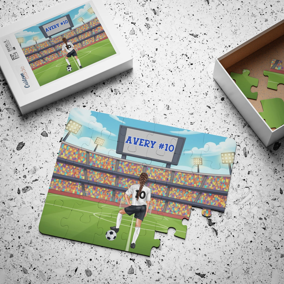 Personalized girls soccer jigsaw puzzle displayed next to its box, showing a stadium soccer scene with a player whose jersey includes a customizable name and number, with loose puzzle pieces scattered inside the box.