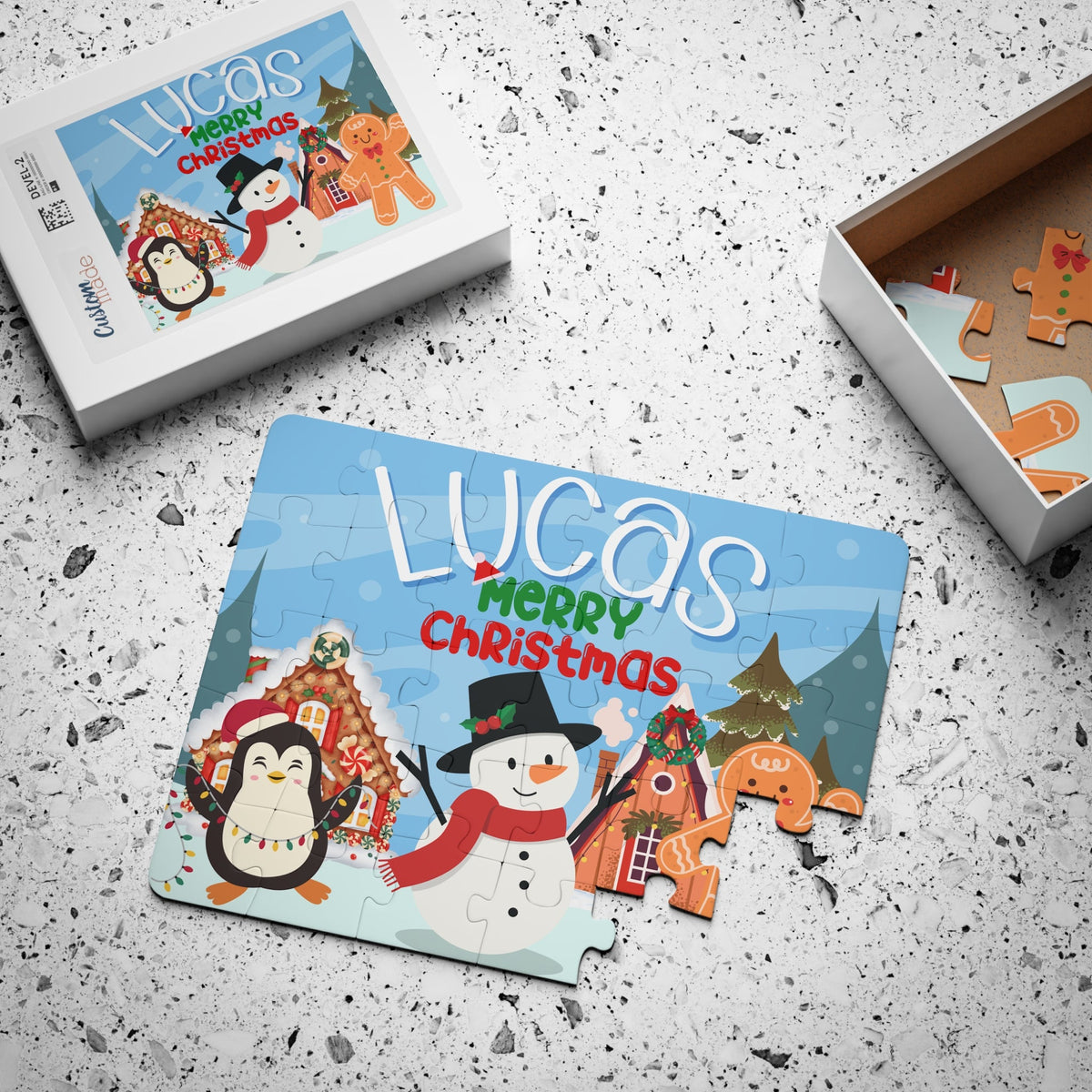 Custom Christmas name jigsaw puzzle for kids displayed with the product box, featuring a snowy holiday scene with a snowman, penguin, gingerbread house, and trees, with the child’s name personalized across the top of the puzzle design.