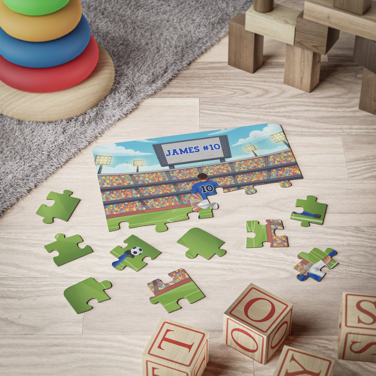 Personalized boys soccer jigsaw puzzle displayed on a playroom floor with scattered jigsaw pieces, showing a stadium background with a customizable name on the scoreboard and a customizable number on the jersey.
