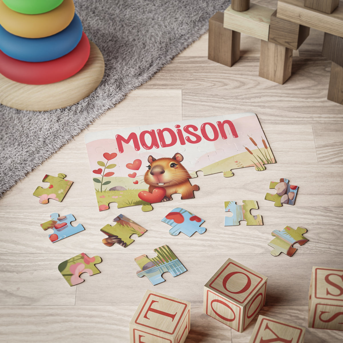 Personalized kids jigsaw puzzle showing a capybara illustration with a customizable name printed across the top of the puzzle, displayed on the floor with scattered jigsaw pieces and children’s letter blocks nearby.