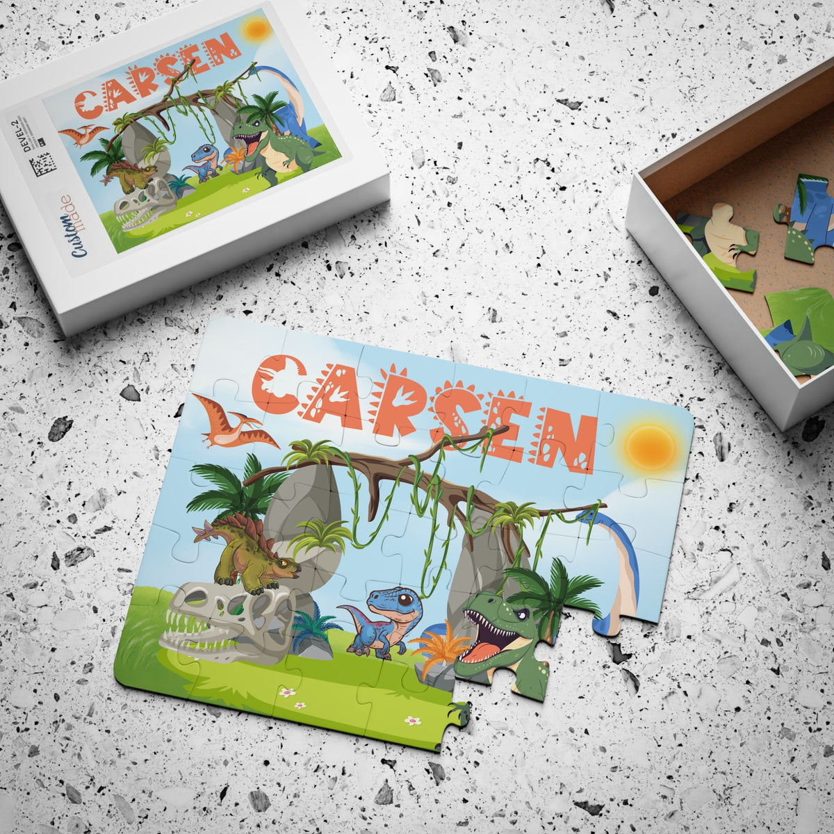Personalized dinosaur jigsaw puzzle for kids with matching puzzle box. Features a bright dinosaur jungle scene with a customizable name printed across the top of the puzzle. The name is chosen by the customer and appears on both the puzzle and the box lid.