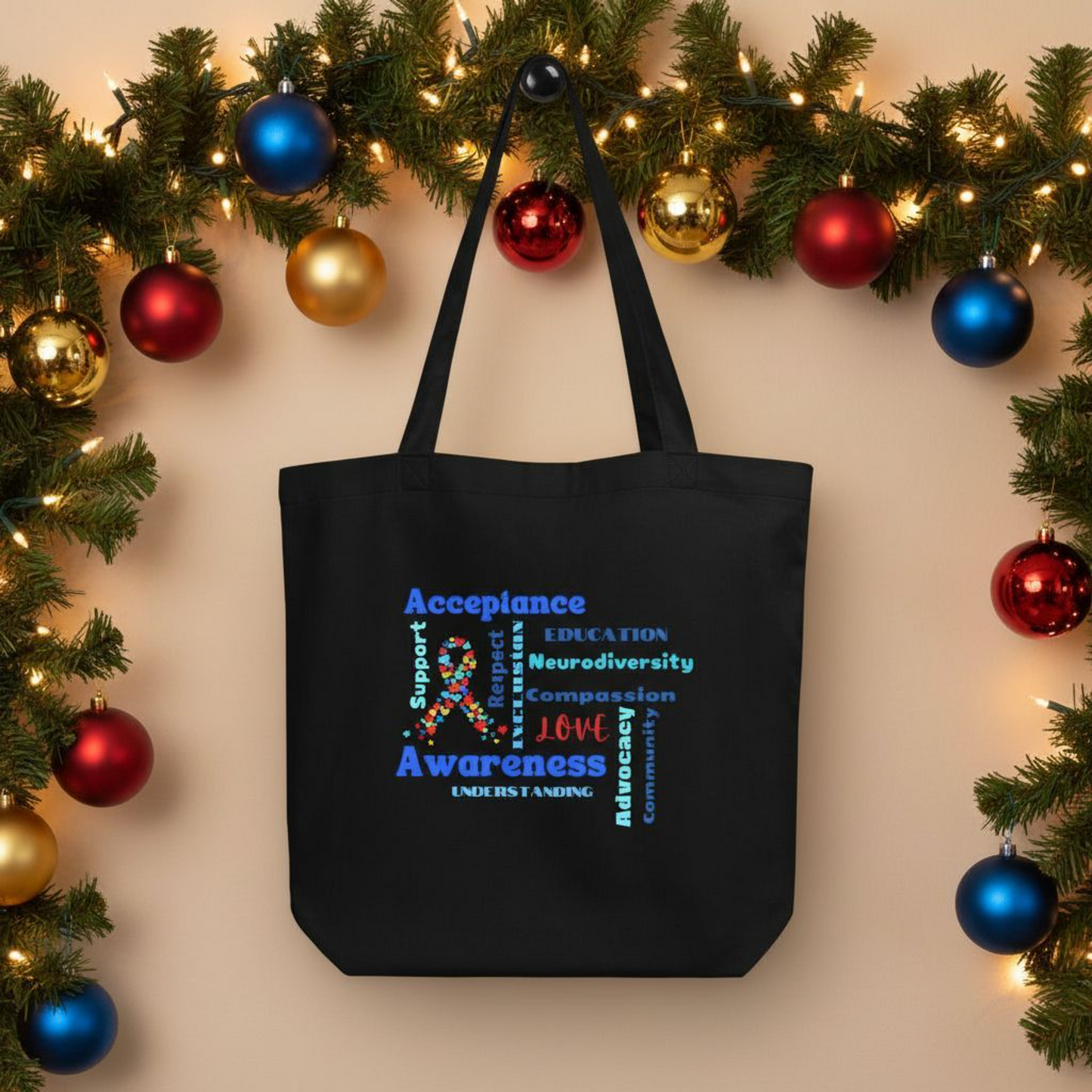 Black eco cotton tote bag displayed with holiday decor, showing an autism acceptance word art design with a multicolor ribbon and words such as acceptance, awareness, education, neurodiversity, compassion, love, advocacy, community, support, respect, inclusion, and understanding.