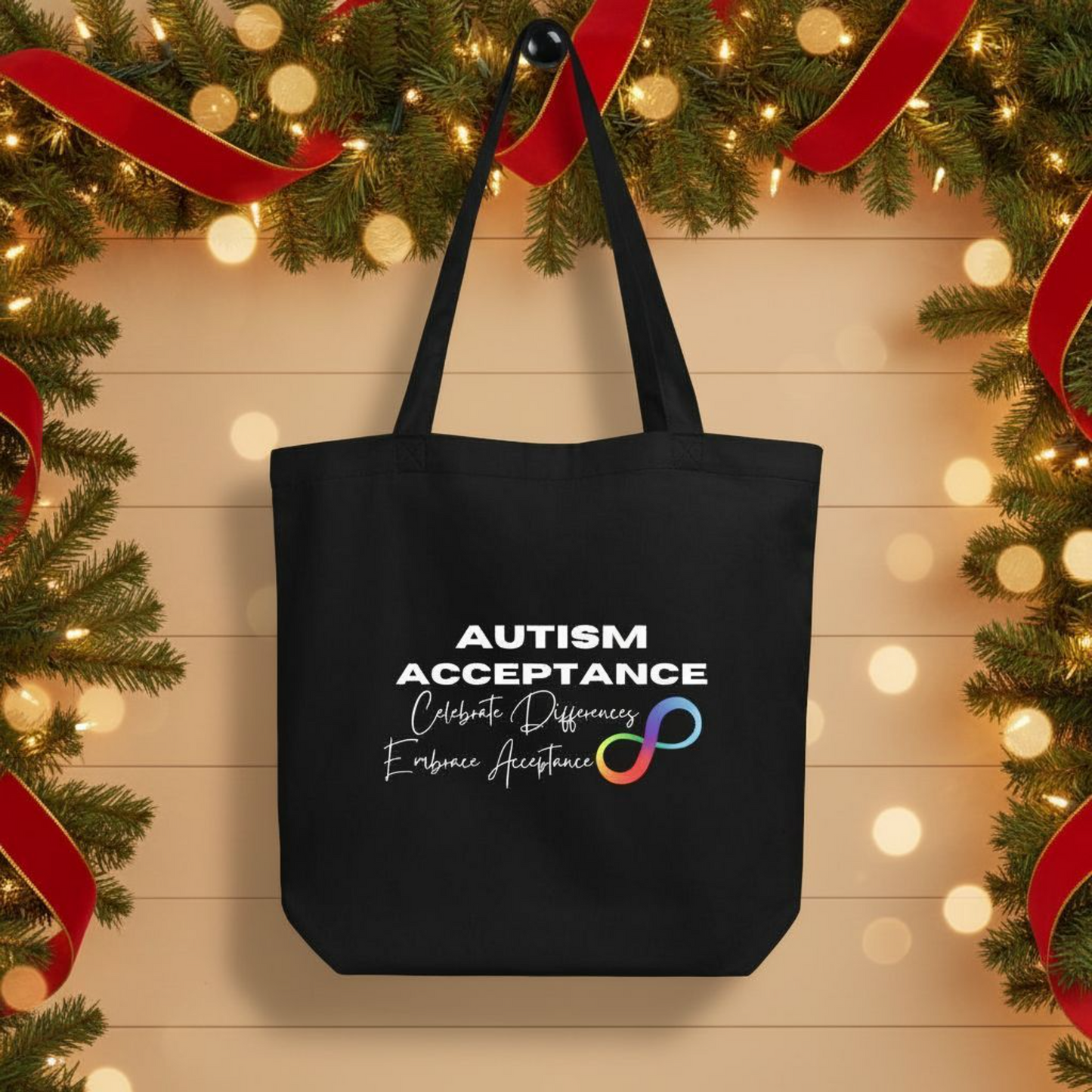 Black eco cotton tote bag hanging in a festive holiday setting with “Autism Acceptance” text, rainbow infinity symbol, and “Celebrate Differences, Embrace Acceptance” script on the front.