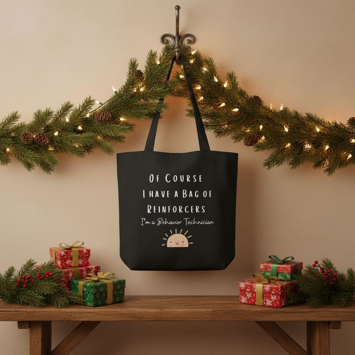 Black eco cotton tote bag hanging under a holiday greenery garland, featuring the full quote “Of course I have a bag of reinforcers, I’m a Behavior Technician” with a smiling sun graphic.