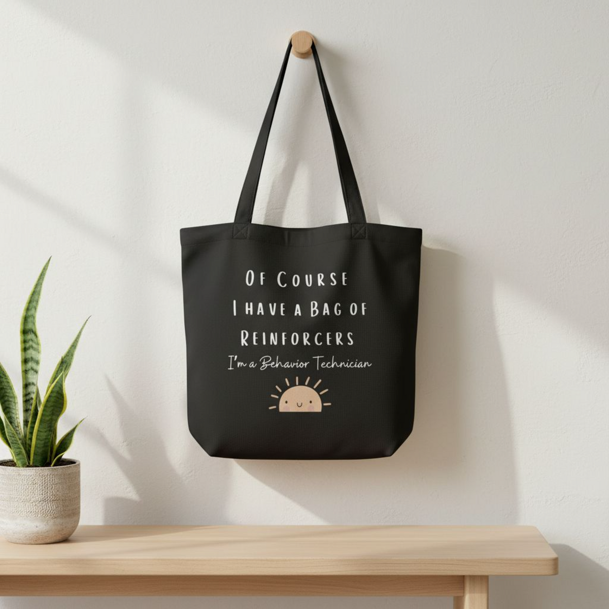 Black eco cotton tote bag hanging on a wall in a minimal setting, displaying the quote “Of course I have a bag of reinforcers, I’m a Behavior Technician” with a small sun illustration.
