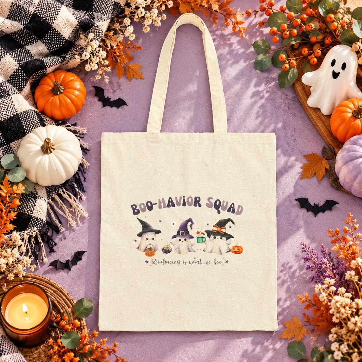 A cream canvas tote bag with a Halloween-themed 'BOO-HAVOR SQUAD' graphic, styled in a flat lay on a lavender background surrounded by autumn decorations including pumpkins, a candle, a ghost, and black bats.