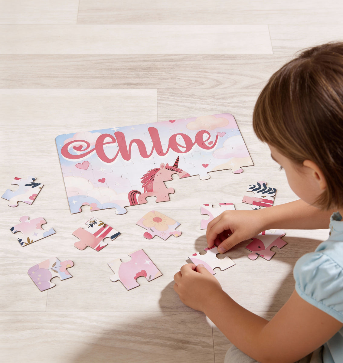 Child playing with a personalized unicorn name puzzle on the floor, placing jigsaw pieces together. The customizable name appears across the top of the puzzle, surrounded by soft pastel artwork and heart details.