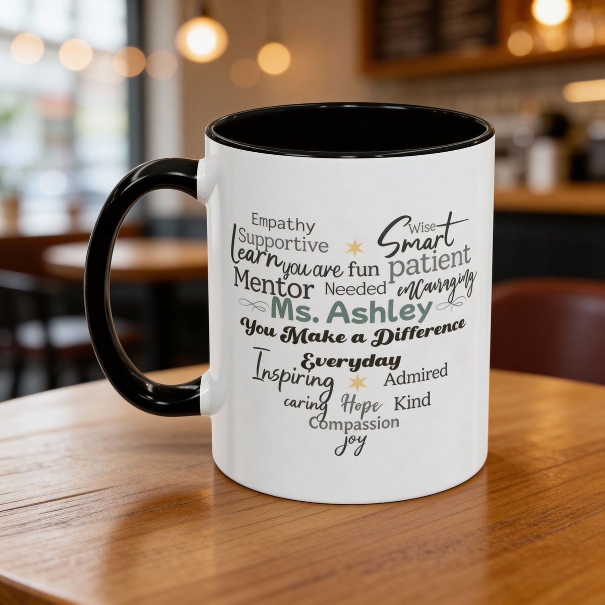 White ceramic accent mug with black handle and interior, featuring a heart-shaped word cloud design with positive traits and a customizable name printed at the center. Mug shown on a wooden table in a café-style setting. Available in 11oz and 15oz sizes.