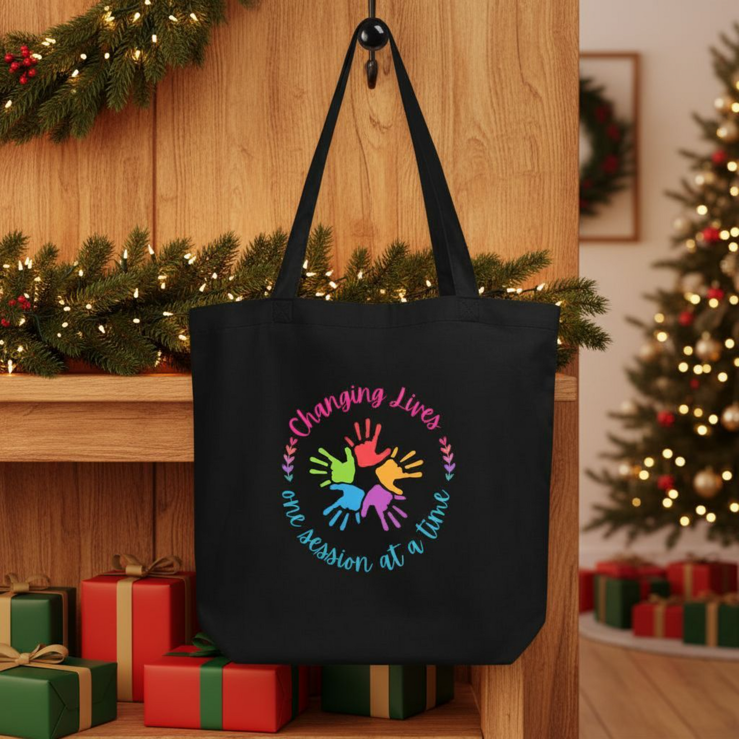 Black eco cotton tote bag displayed in a festive setting, showing a rainbow handprint circle graphic and the words “Changing Lives One Session at a Time.”