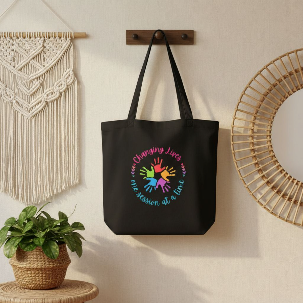 Black eco cotton tote bag hanging on a wall hook, featuring a colorful handprint circle design with the phrase “Changing Lives One Session at a Time.”