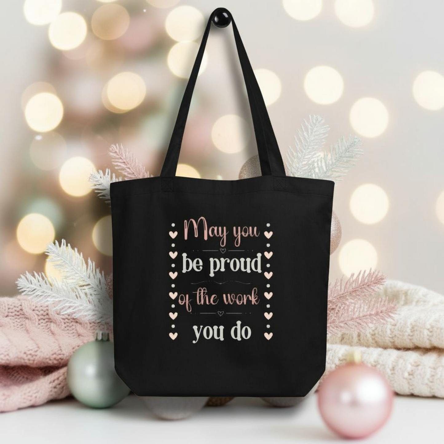 Black eco tote bag displayed in a cozy holiday setting with the quote “May you be proud of the work you do,” printed vertically with decorative hearts.