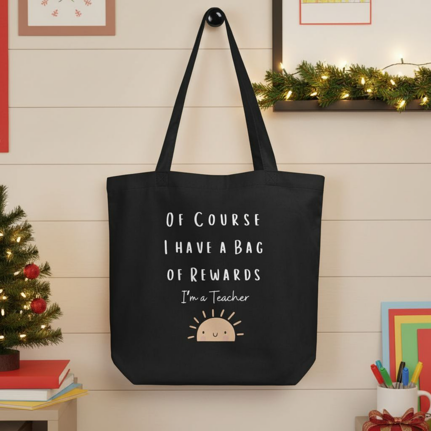 Reusable black eco cotton tote bag displayed in a Holiday classroom setting, showing the phrase “Of course I have a bag of rewards, I’m a teacher” with a minimalist sun graphic, intended as a funny teacher gift tote.
