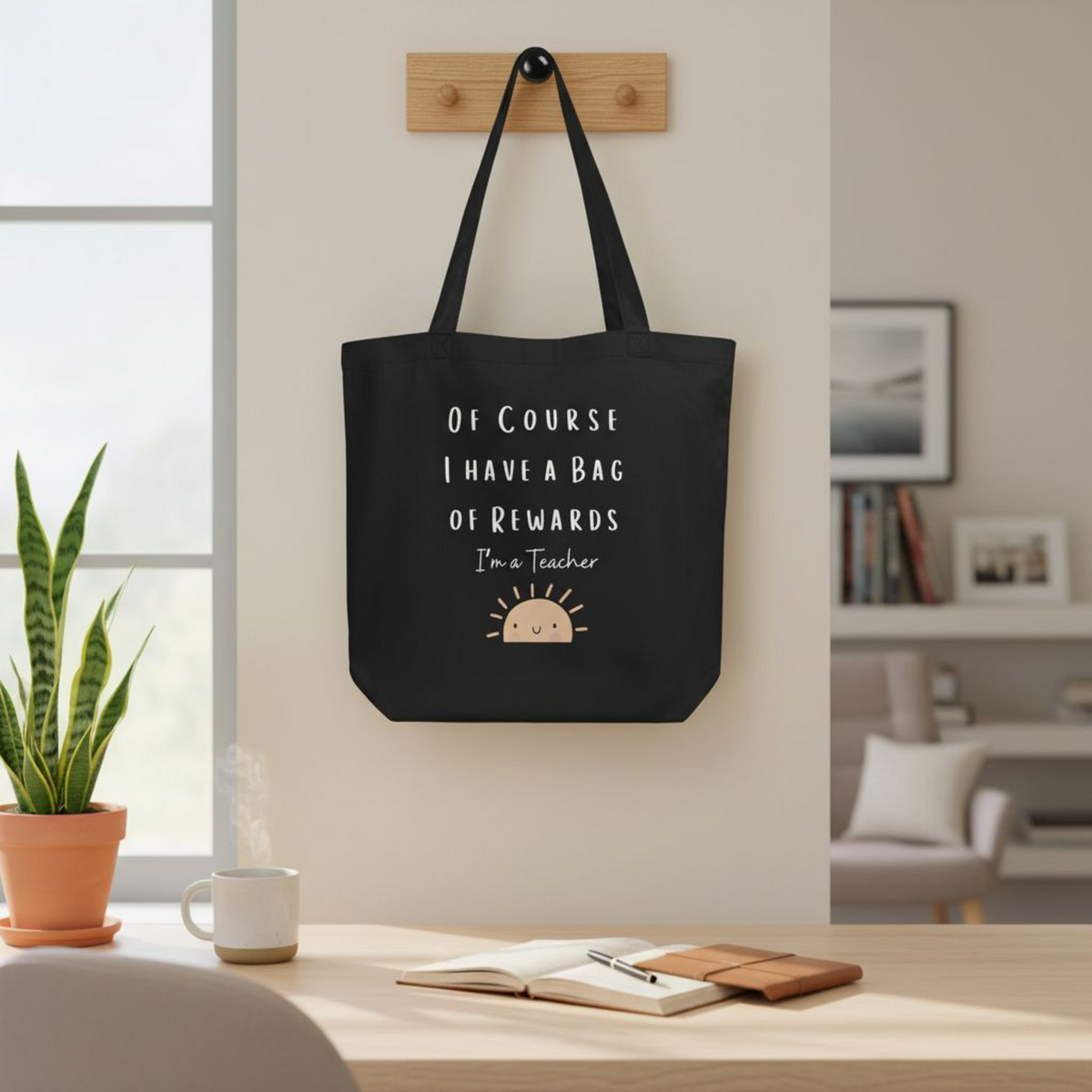 Black eco cotton tote bag hanging on a wall, featuring the quote “Of course I have a bag of rewards, I’m a teacher” with a small smiling sun illustration, designed as a reusable teacher tote bag.
