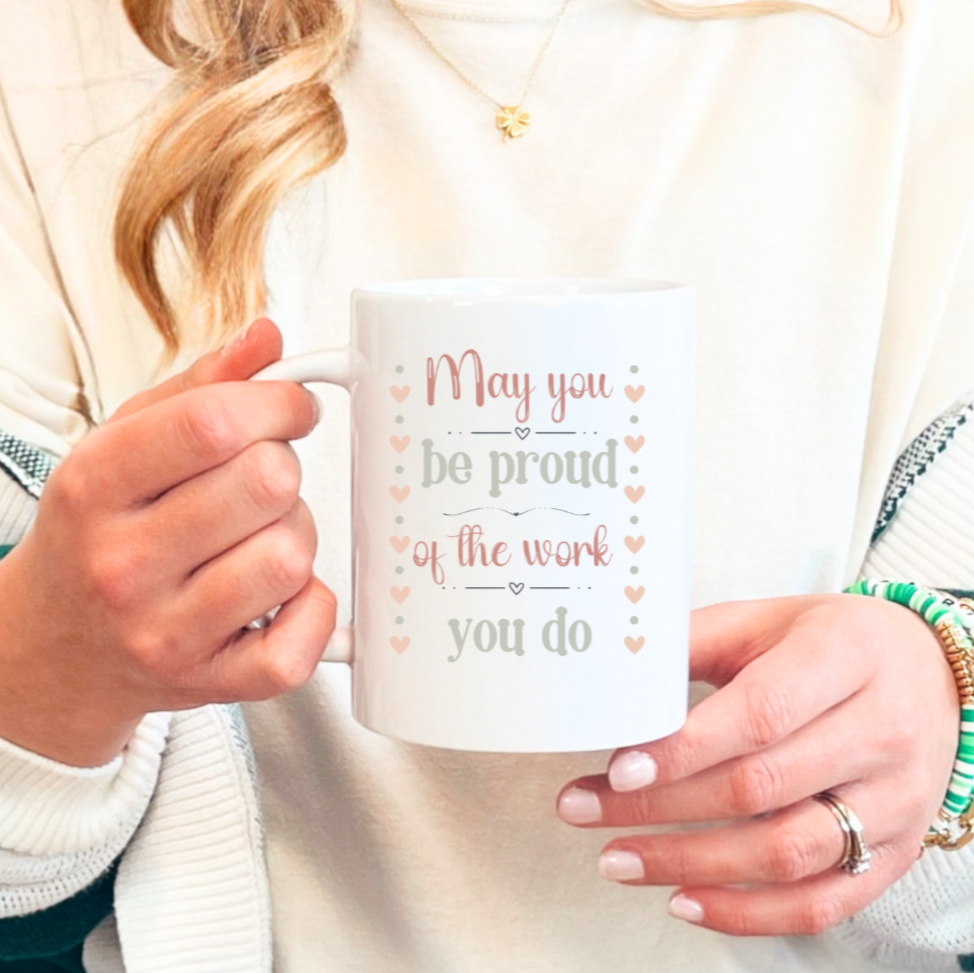 Lifestyle photo of a person holding a personalized white ceramic mug with the phrase “May you be proud of the work you do,” showing a customizable name printed on the side of the mug in a cozy indoor setting.