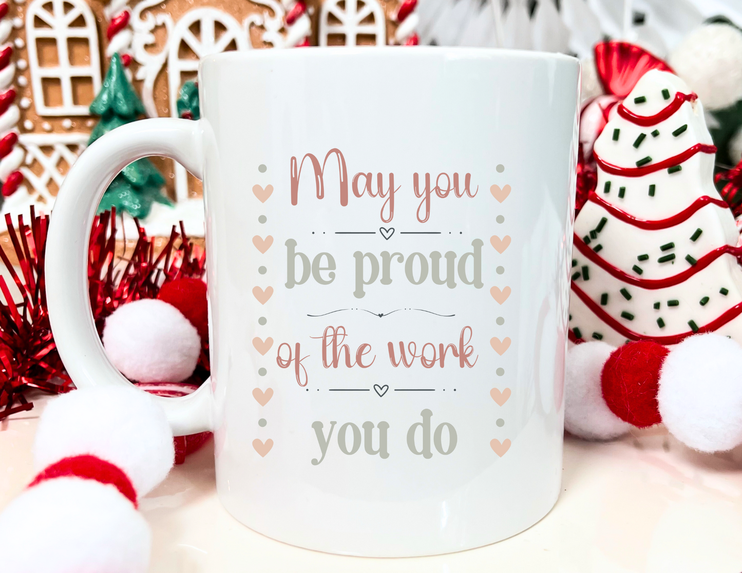 Personalized white ceramic mug featuring the message “May you be proud of the work you do,” with a customizable name printed on the side of the mug, styled with festive holiday decorations including gingerbread houses and ornaments.