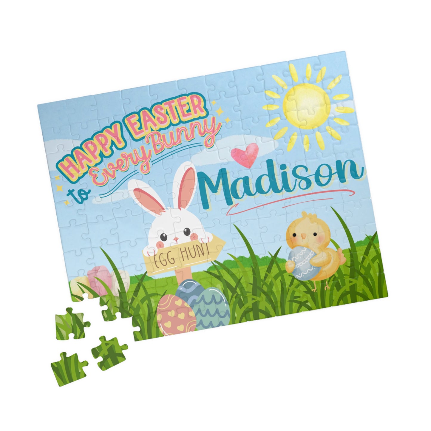 Personalized Easter bunny 110-piece jigsaw puzzle shown at an angle with a few loose jigsaw pieces nearby. The spring illustration includes eggs, grass, and animals, with a custom name integrated into the artwork.