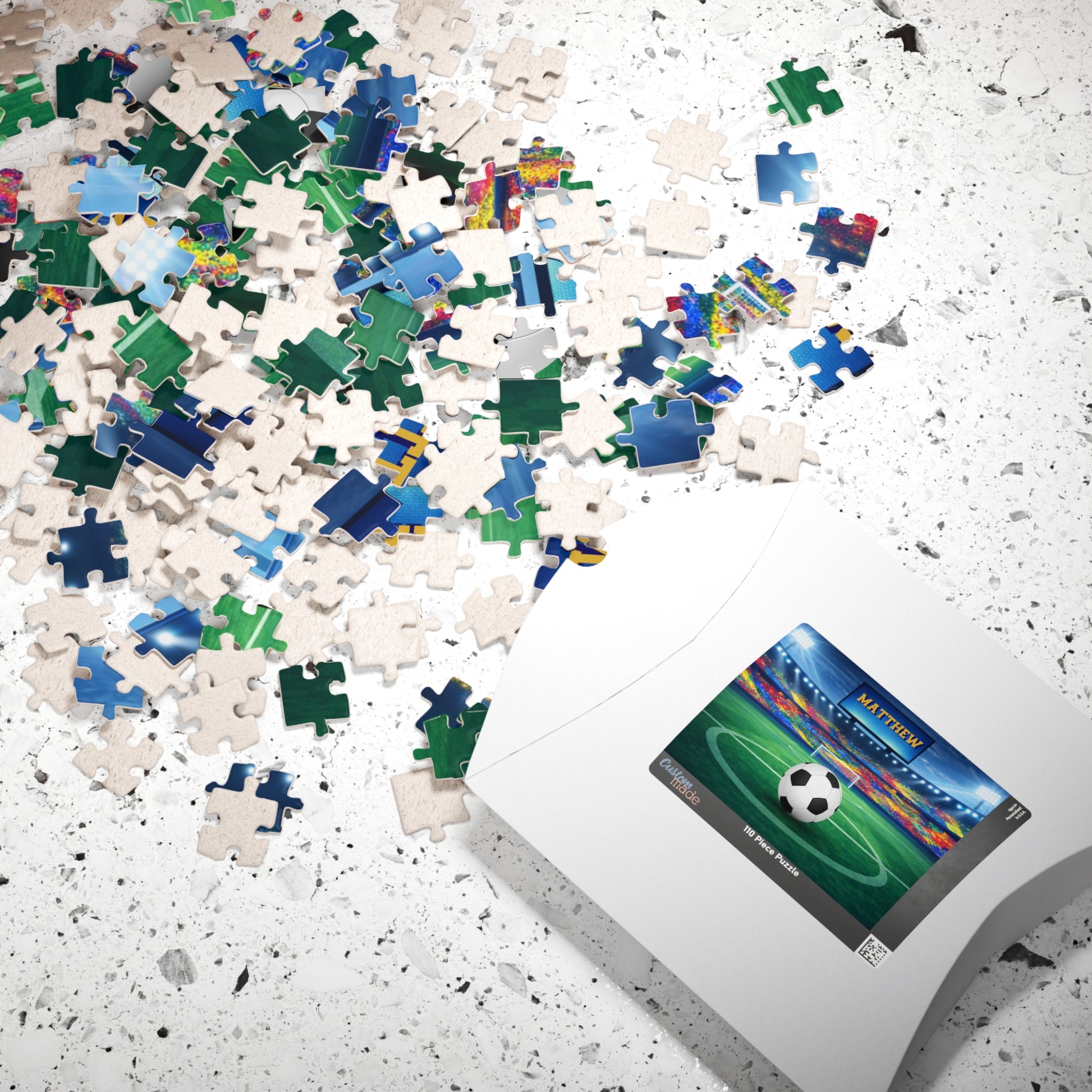 Personalized soccer stadium jigsaw puzzle shown with loose jigsaw pieces spread out next to the puzzle box. Stadium scene includes a customizable name on the jumbo screen and a customizable soccer ball. 110-piece puzzle.