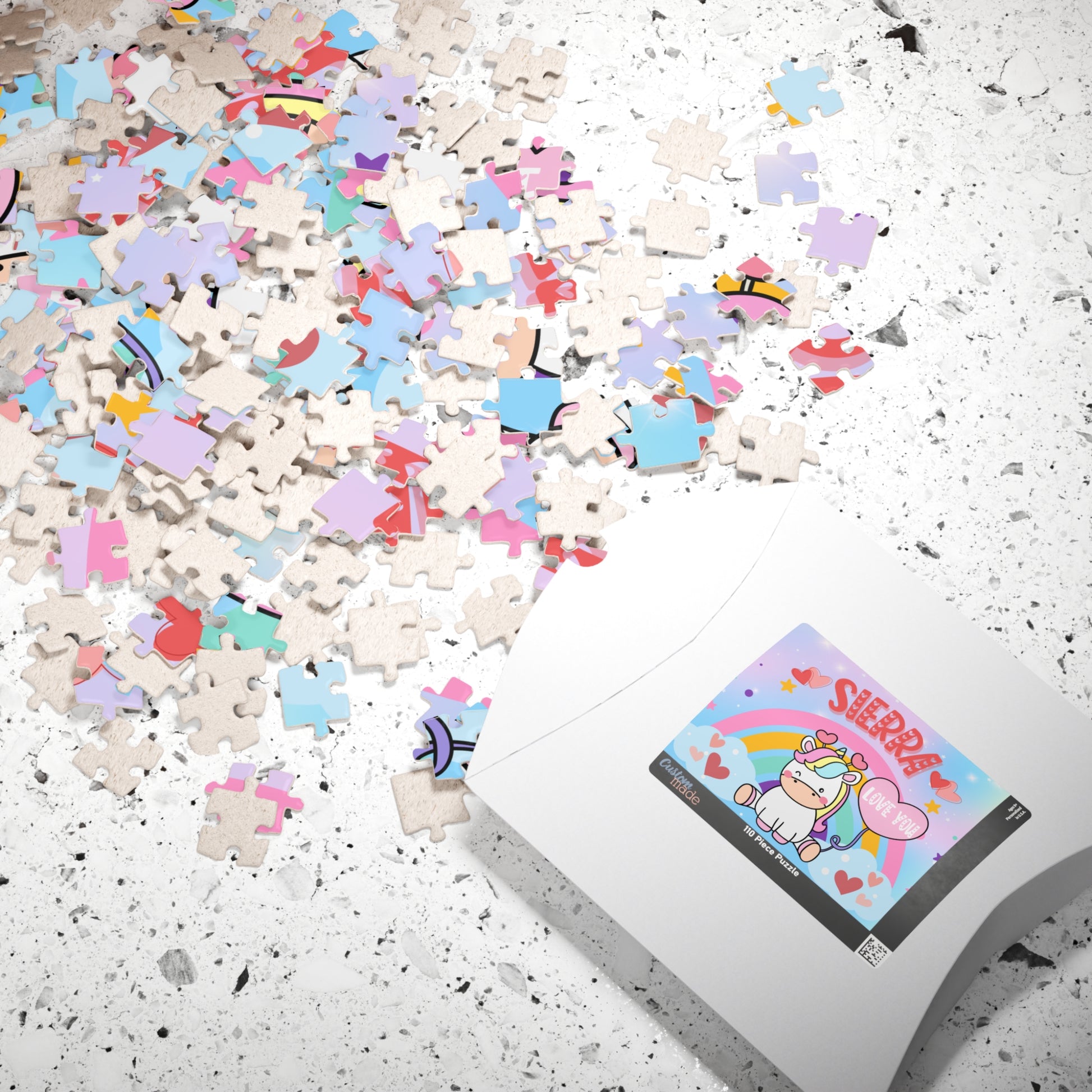 Scattered 110-piece unicorn rainbow jigsaw puzzle pieces on a countertop with packaging box showing the customizable child’s name on the label.