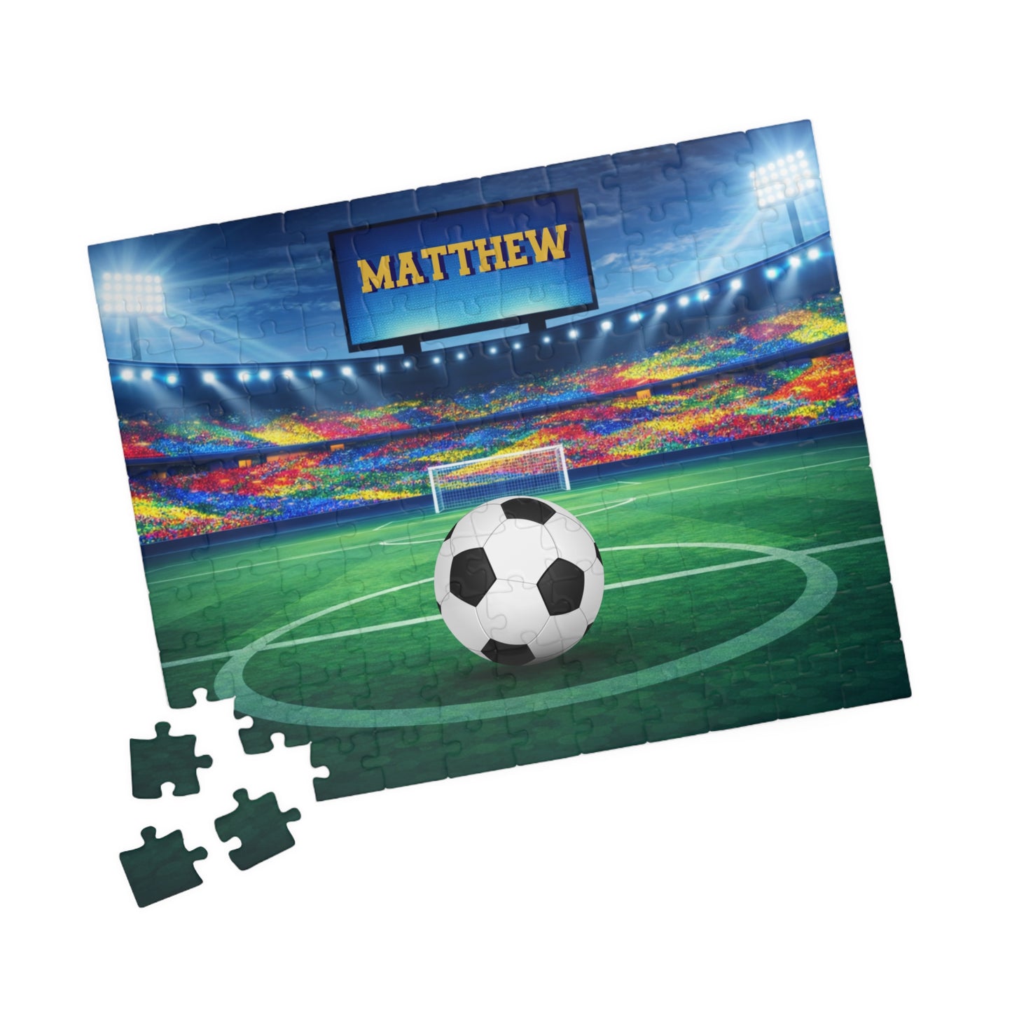 Personalized soccer stadium jigsaw puzzle shown at an angle with several loose jigsaw pieces nearby. Customizable name appears on the stadium screen, and the soccer ball design is customizable. 110-piece puzzle for older kids and families
