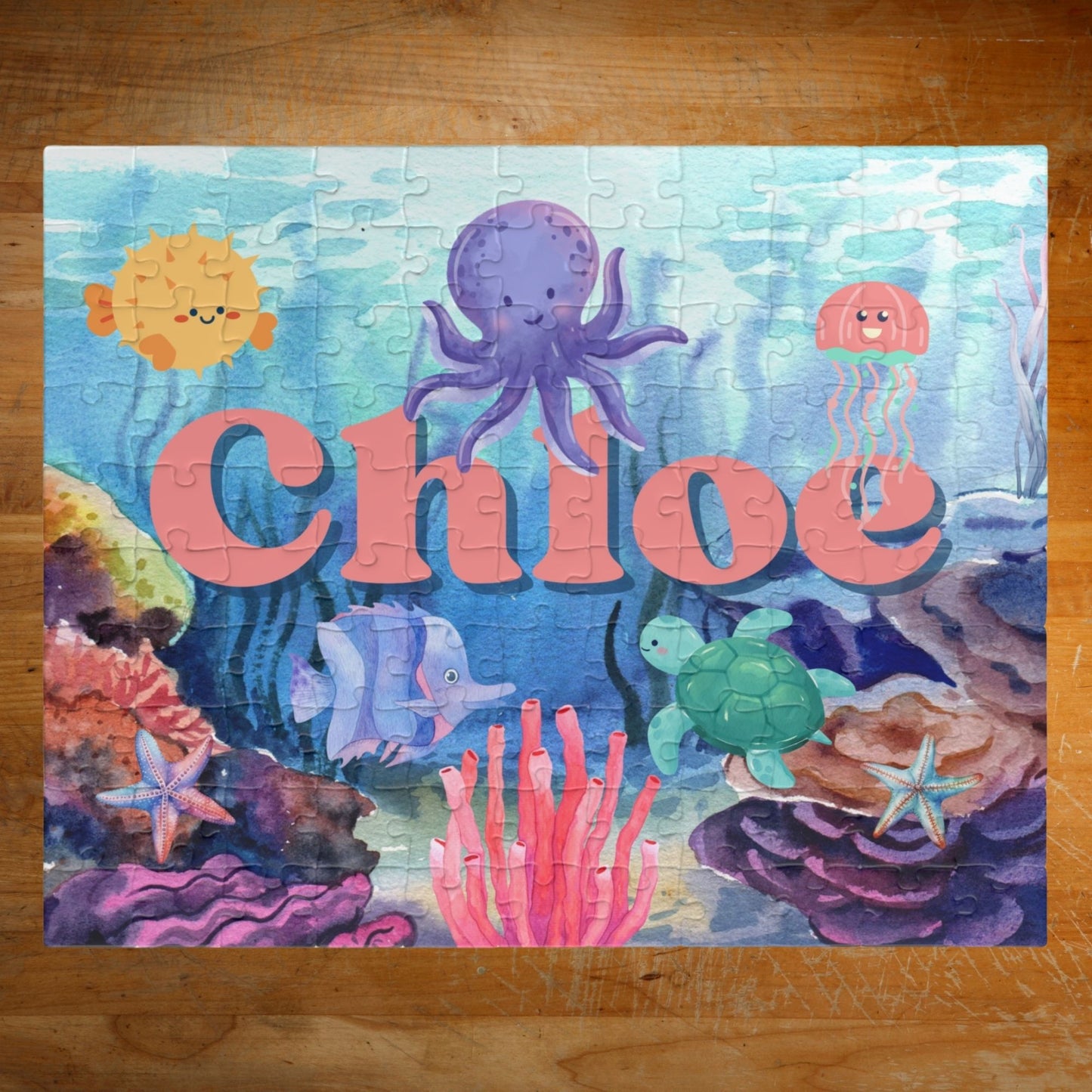 Personalized ocean-themed 110-piece jigsaw puzzle featuring a colorful underwater scene with sea animals, coral, and fish. A customizable child’s name appears prominently across the center of the puzzle design.