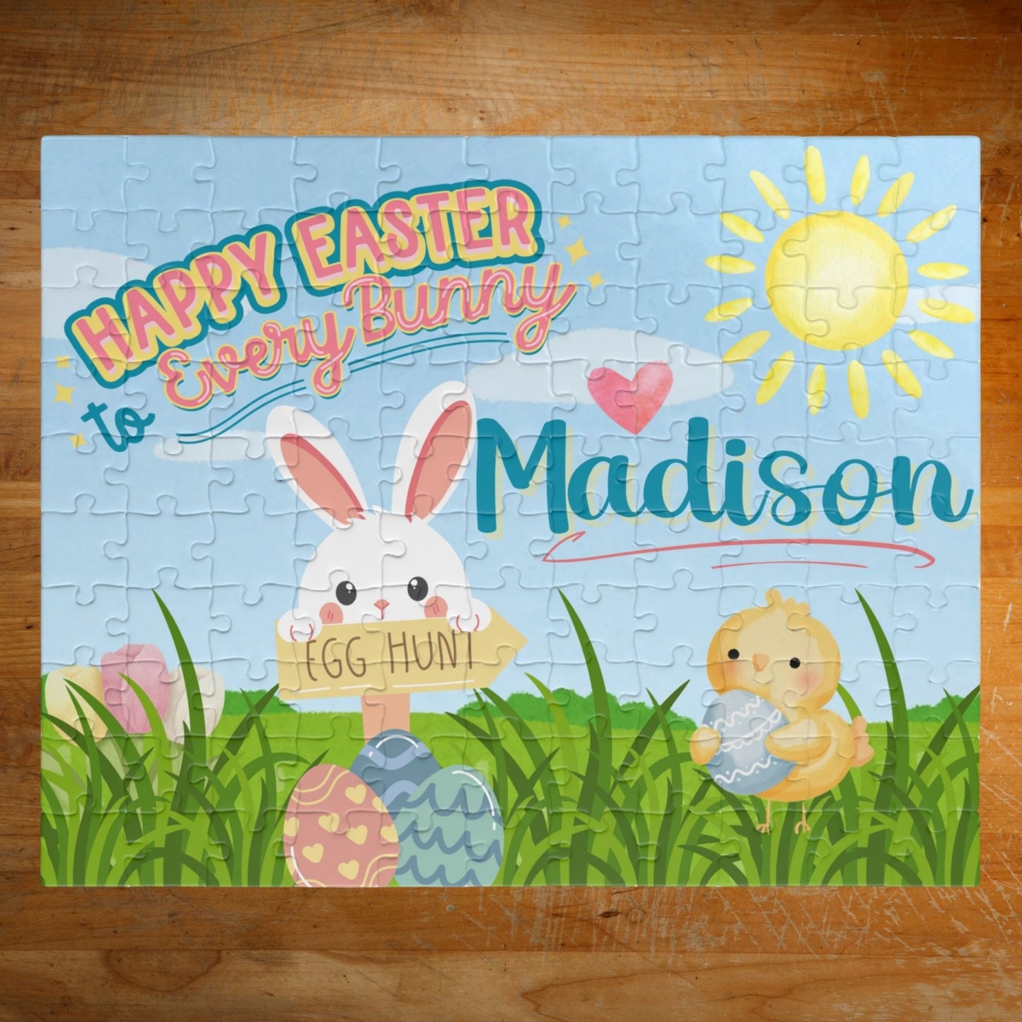 Personalized Easter-themed 110-piece jigsaw puzzle featuring a spring scene with a bunny, chick, pastel eggs, green grass, and sunshine. A customizable name appears across the center of the puzzle design.