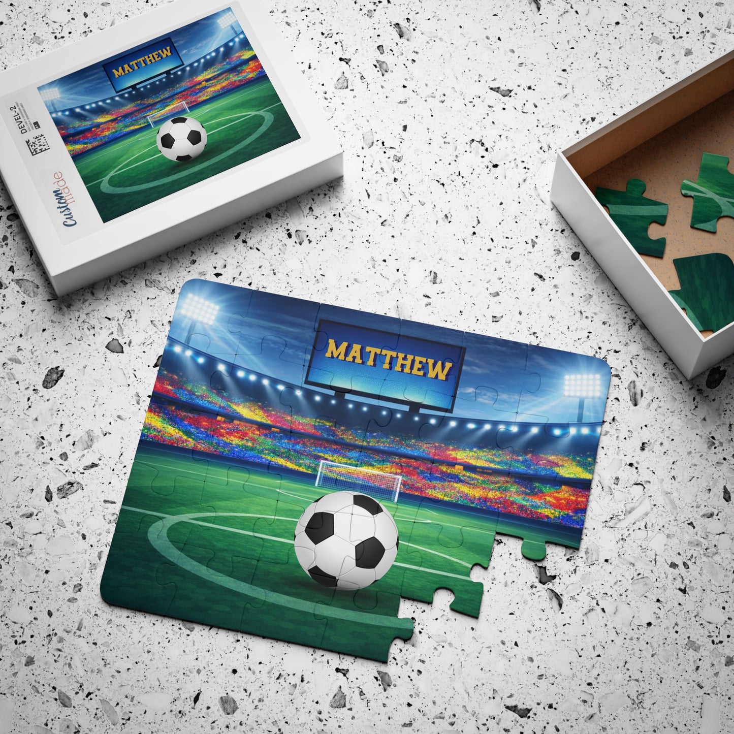 Personalized soccer jigsaw puzzle shown partially assembled with loose jigsaw pieces and gift box packaging, featuring a stadium background and a customizable name printed on the scoreboard above the goal.