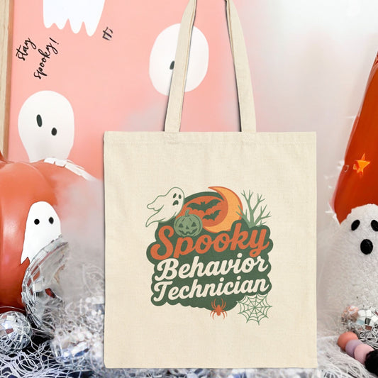 Natural canvas tote bag featuring a Halloween-themed design that reads “Spooky Behavior Technician,” with a ghost, pumpkin, crescent moon, bats, spider, and spiderweb, styled in warm fall colors and displayed against a festive Halloween background.
