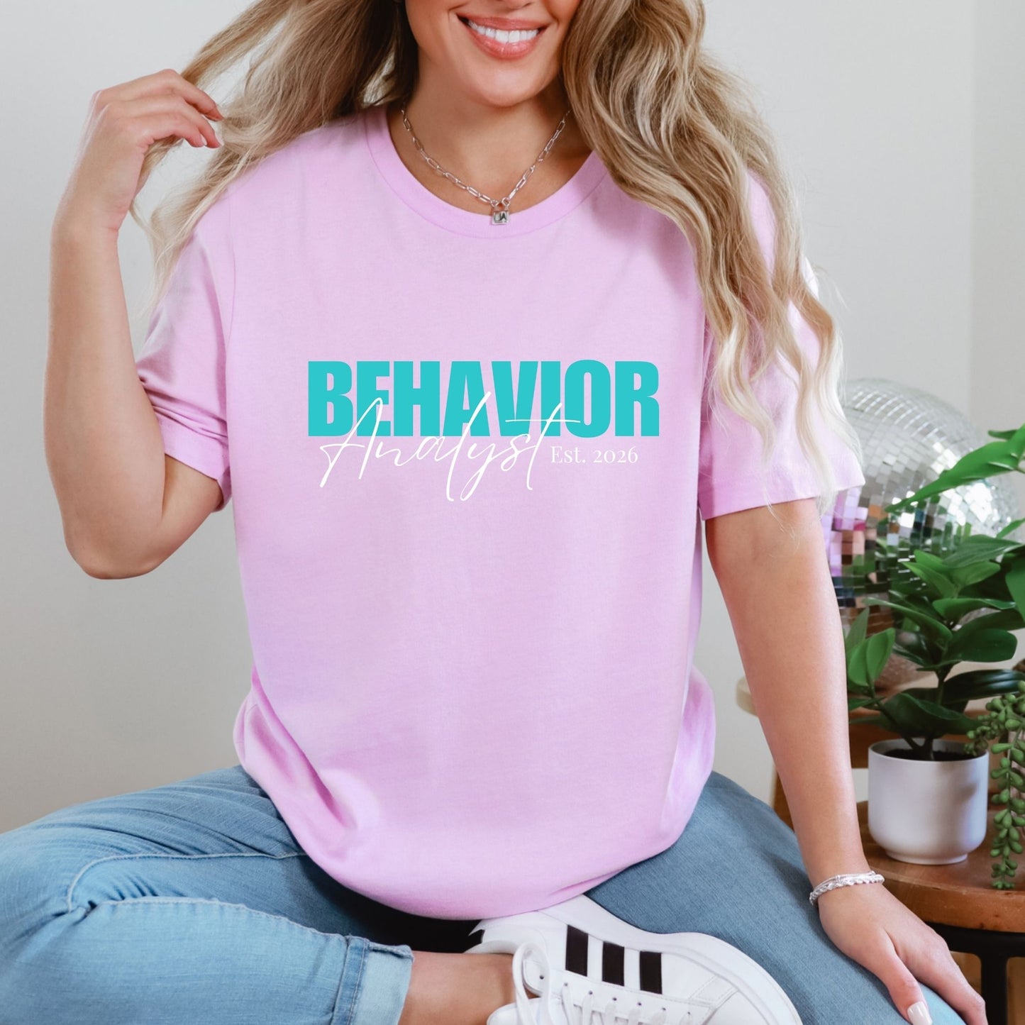 Model wearing a lilac t-shirt with a modern teal and white “Behavior Analyst” script design featuring “Est. 2026” on the front; shirt styled casually with jeans and sneakers. Design available in multiple color options.