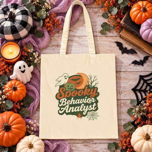 Natural canvas tote bag featuring a Halloween-themed design that reads “Spooky Behavior Analyst,” with a ghost, pumpkin, crescent moon, bats, spider, and web, displayed on a festive fall background with pumpkins.