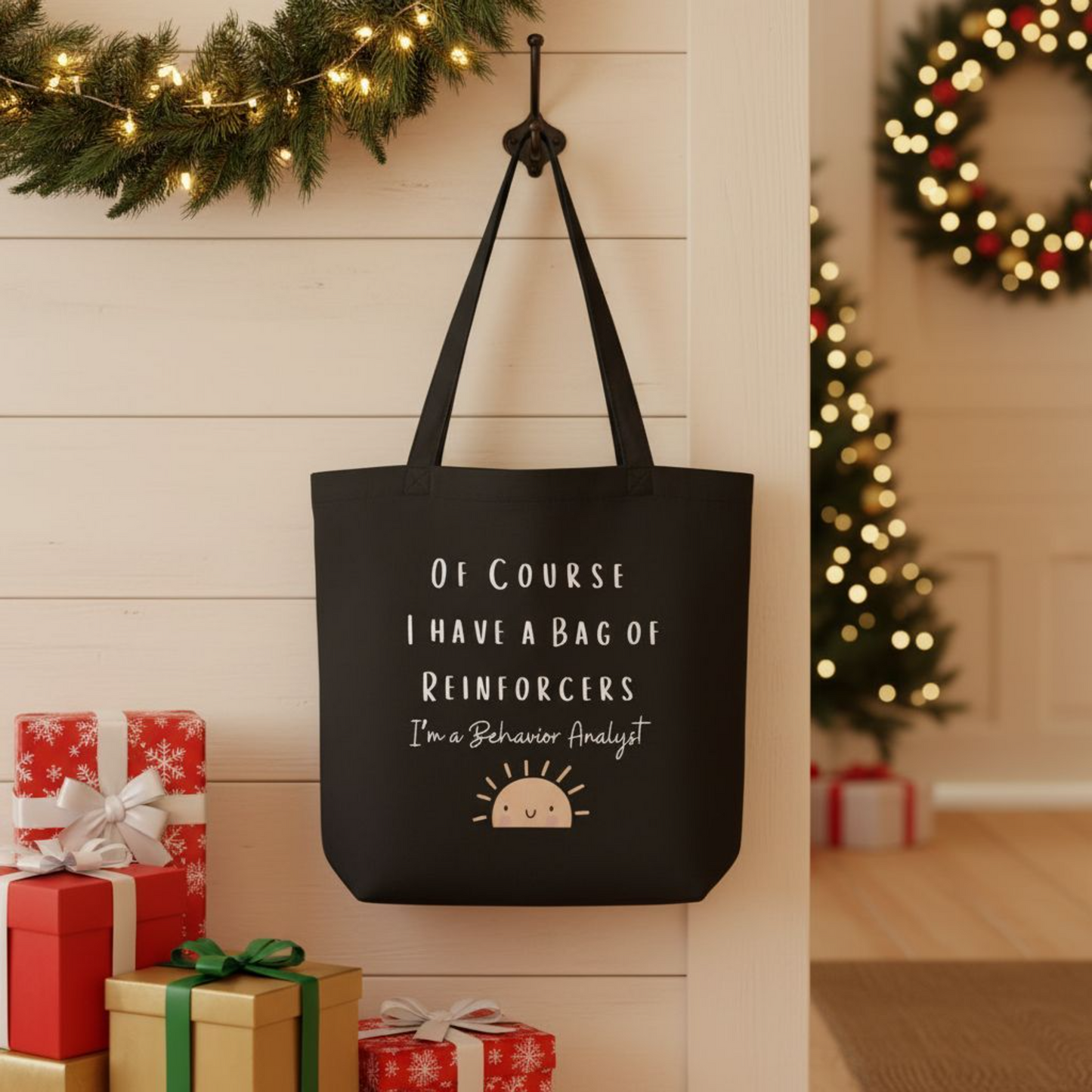 Black eco cotton tote bag displayed in a holiday setting with gift boxes, showing white lettering “Of Course I Have a Bag of Reinforcers – I’m a Behavior Analyst” and a smiling sun design, reusable tote for behavior analysts.